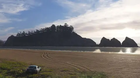 Carro verde conversível numa praia de areia, com ilha rochosa coberta de pinheiros e mar ao fundo, em cena de jogo