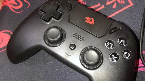 Controle de videogame preto Redragon com analógicos e botões frontais, sobre mousepad vermelho e preto